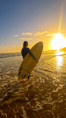LENS FLARE: Female surfer in wetsuit and with wooden surfboard walks through shallows at sunset. Setting sun casts warm glow on water, as she returns from an evening surf session at Moroccan beach.