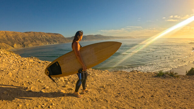 LENS FLARE, PORTRAIT: Woman in a wetsuit holding a surfboard walks along a scenic cliff path overlooking the famous Moroccan surf break. She goes for an evening surfing in beautiful golden light.