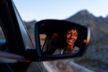 Rearview mirror reflection of a smiling woman during a sunset scenic drive in the Sierra de Tramuntana. Concept of travel, freedom, automotive lifestyle.