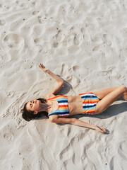 Sunlit woman lying on white sandy beach, wearing a bright striped bikini. Relaxed pose, arms at...