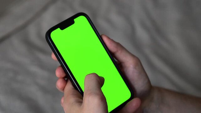 Male hand holding modern smartphone with blank green display on bed background.
Blank green screen on mobile phone.
Background replacement for design.
Modern technologies and video editing