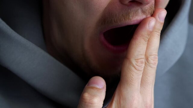 A man covers his mouth with his hand when he yawns.
A tired man yawns in a room.
A man opens his mouth wide when he yawns and covers it with his palm.
A sleepy and tired man