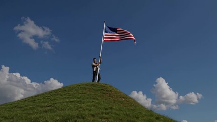 A man stands atop a grassy hill, holding an American flag against a blue sky. The low-angle shot gives a cinematic video feel, emphasizing patriotism. - Powered by Adobe