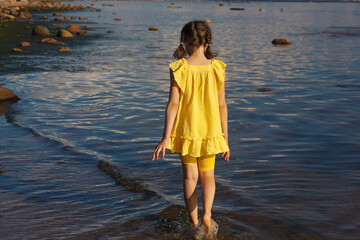 Little girl walking along seashore in yellow dress on sunny day, view from back