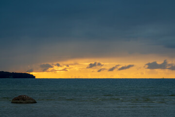 Dramatic ocean sunset with golden sky and dark storm clouds over water. Beautiful seascape showing moody evening light with rocky coastline and calm sea surface
