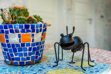 On the table, there is a cactus plant in a mosaic-patterned pot and next to it is a cartoonish ant figure made of black metal.