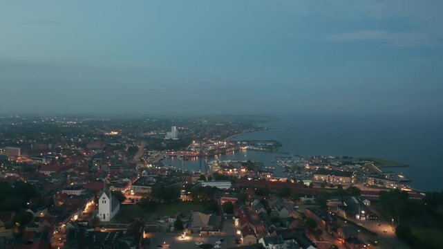 Aerial night view of Thisted, Denmark. Town lights, marina, Limfjord coastline, and surrounding neighborhoods create a tranquil Nordic cityscape under a twilight sky
