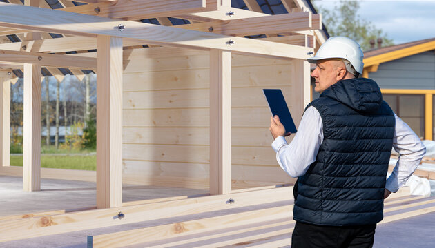 Construction. Engineer or architect in hard hat inspecting wooden house frame with tablet, building site supervision, architecture, project management, residential development.