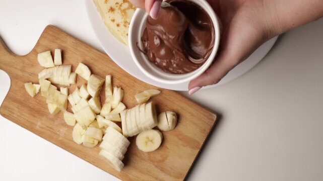 stirring chocolate spread in small white bowl with spoon. close-up of sweet filling preparation next to sliced bananas on wooden cutting board for homemade dessert