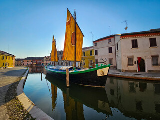 Traditional sailboat with orange and yellow sails docked in a canal in Comacchio, Italy, with historic buildings