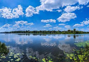 Clouds are reflected in the river. Puffs of steam hover over the surface of the water. Landscape with sky and waves. Freshwater reserves. Water lilies overgrow the surface. Day blue sky.