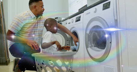 Kneeling father-son duo loading clothes from plastic basket into washer in laundry room, copy space