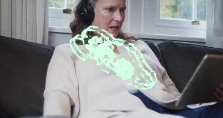 Reclining woman wearing cream cardigan and headphones on sofa at home, holding tablet by window
