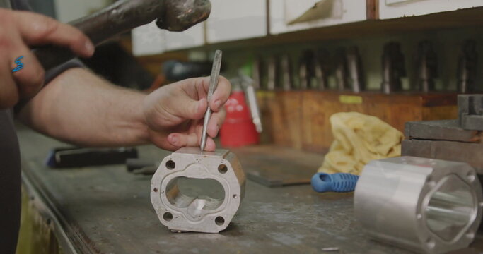 Striking machined aluminum block on metal workshop bench by worker's hands, with hammer and chisel