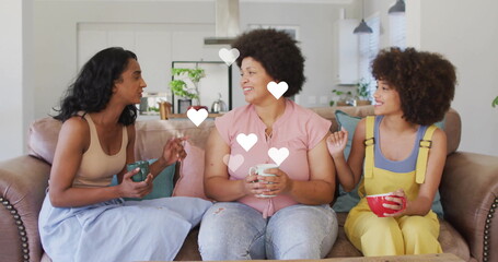 Smiling three women leaning toward each other and chatting on leather sofa at home, with mugs