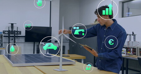Male engineer holding tablet, adjusting wind turbine and solar panel prototype at lab, with metrics