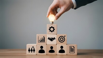 Hand placing a light bulb atop a pyramid of blocks with business icons, symbolizing innovation and strategy.