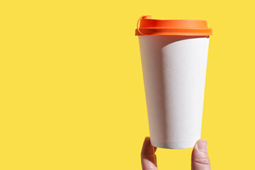 Hand holding white paper cup with orange lid against bright yellow background for conceptual design.