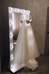 Wedding photo of part, element of white lace dress hanging on hanger against mirror with lamps in hotel room in morning.