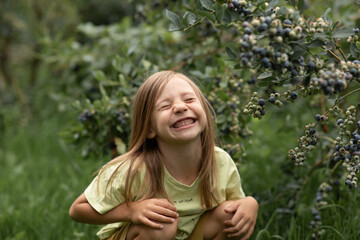 a close-up portrait of a blonde girl sitting next to green bushes, smiling broadly with her eyes closed
