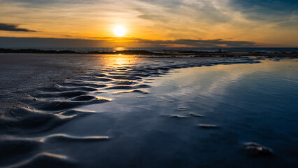Beach water detail with repetitive sand arc formation and colorful sunset in background