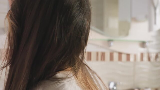 Back view young lady with long hair in well lit tiled bathroom stretches toward shelf to place item, soft highlights on hair and tiles, mirror and sink blur in background, everyday hygiene action
