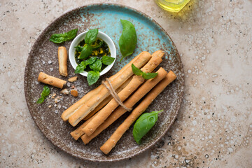Brown plate with salted breadsticks grissini and basil oil, horizontal shot on a beige granite background, flat lay