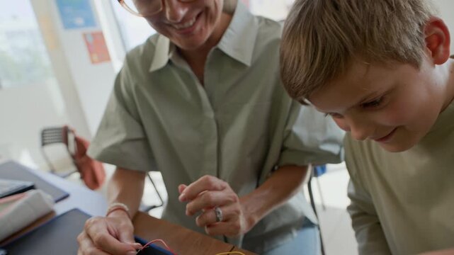 Tilt up of young female teacher and clever schoolboy connecting colorful wires to circuit board on desk while carrying out technical assignment at lesson
