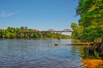 A serene Coosa river flows gently under a clear blue sky, while historic Wetumpka bridge arches gracefully overhead. Lush greenery lines the banks, enhancing the peaceful atmosphere on this sunny day.