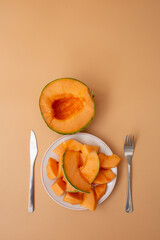 Half a cantaloupe and a plate with sliced melon pieces, served with fork and knife on a pastel background.