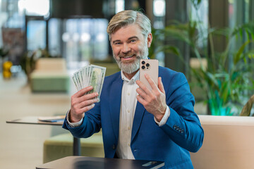 Successful happy rich mature businessman holding smartphone and counting dollar bill money fan...