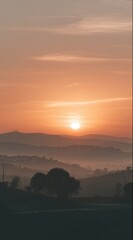 Obraz premium Sunset over hazy hills with layers of silhouettes and warm tones. The sky is orange and the sun is a bright orb. Fog drapes softly in the valleys