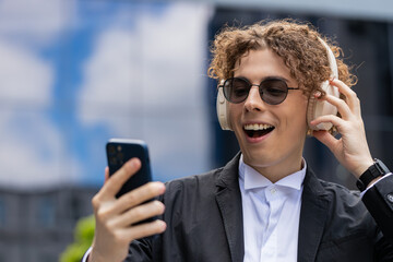 Happy joyful Caucasian manager listens to music on wireless headphones while walking on the street...