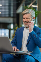 Confident mature businessman in suit answering to client on mobile phone call, using smartphone and laptop. Conversation. Caucasian bearded man guy working online job, talking in modern office lobby.