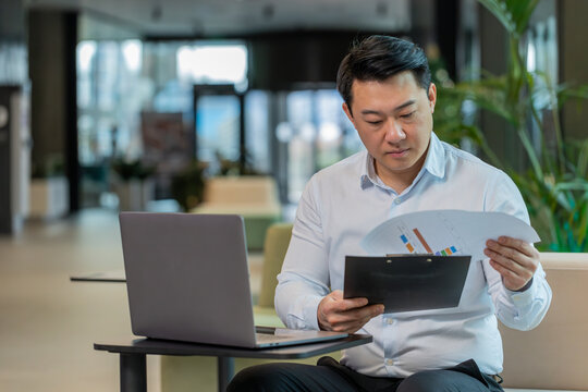 Chinese businessman working with documents, analyzing information, reading papers prepare financial report. Young male entrepreneur in formal using laptop analyzing graph documents in office lobby.