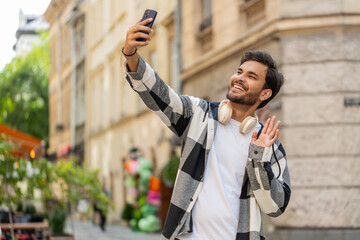 Cheerful Indian man blogger taking selfie on smartphone, communicating video call online with family friends, recording stories for social media vlog, waving hello. Hispanic guy in urban city street.