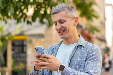 Happy smiling middle-aged man using smartphone typing text messages browsing internet social media web app working chatting online outdoor. Mature gray-haired guy tourist standing in urban city street