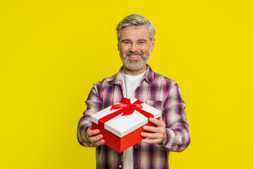 Mature man hands over festive gift box with bright red ribbon, radiating great delight excitement. Middle-aged guy on yellow background reveals joyful mood, celebrating fun occasion with wide smile.