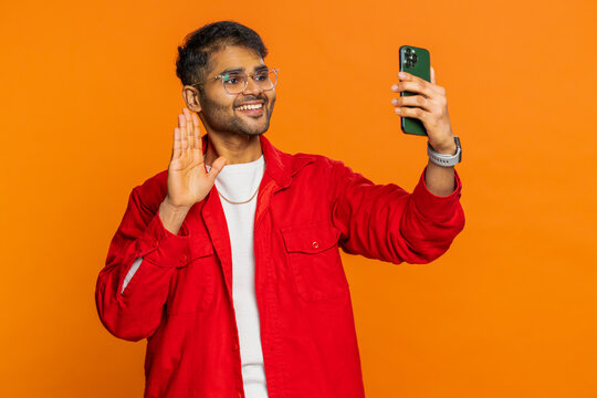 Happy Indian man blogger taking portrait selfie on smartphone, streaming, vlog, communicating record video call online with social media subscribers. Arabian young guy isolated on orange background