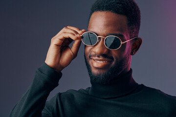 Obraz premium Confident young Black man wearing sunglasses and a turtleneck, smiling against a dark, subtly lit background, representing style and modern fashion