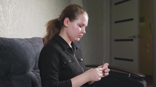 Side view of focused student with neat manicure seated on sofa, gaze fixed on threading needle with steady hands, dark zip jacket, calm concentration, home craft scene, patient practice