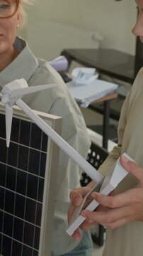 Vertical shot of schoolboy blowing at turbine of windmill mockup while teacher with sollar panel helping him during presentation of usage of renewable sources of energy