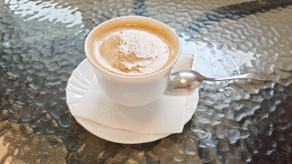 There is a ceramic coffee cup, a saucer with a napkin and a metal spoon on the glass top of the table. Time for a coffee