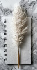 A blank spiral-bound notebook sits on a marble surface,  with a fluffy white pampas grass stem placed diagonally