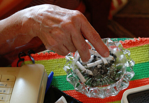 Mano apagando cigarro de tabaco negro con nicotina tras fumar en cenicero de cristal junto a otros pitillos contra ley antitabaco