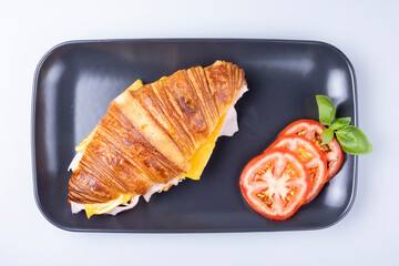 Top view of a ham and cheese croissant sandwich on a black plate with fresh tomato slices and basil. Savory breakfast or lunch meal, perfect for café, menu, or food concept.