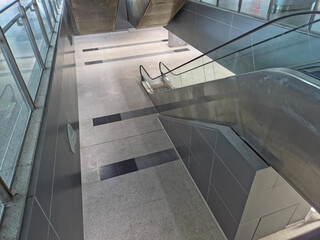 ChatGPT said:

High-angle view of an empty modern passageway, featuring a staircase and escalator descending to a lower level, with sleek gray and black floor tiles and glass-and-metal railings.