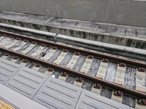 close-up, diagonal view of a modern train track. The parallel metal rails are secured to concrete sleepers with red and black fasteners.