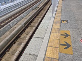 Fototapeta premium Low-angle diagonal view of train tracks and platform edge, featuring a yellow tactile strip with directional arrows, highlighting the functional design of an urban transit area.