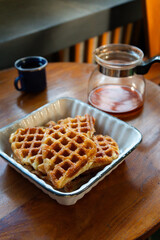 Close-up of freshly baked golden brown croffles served in a ceramic dish on a wooden table. A glass coffee pot and a blue enamel mug complete the cozy breakfast setup.